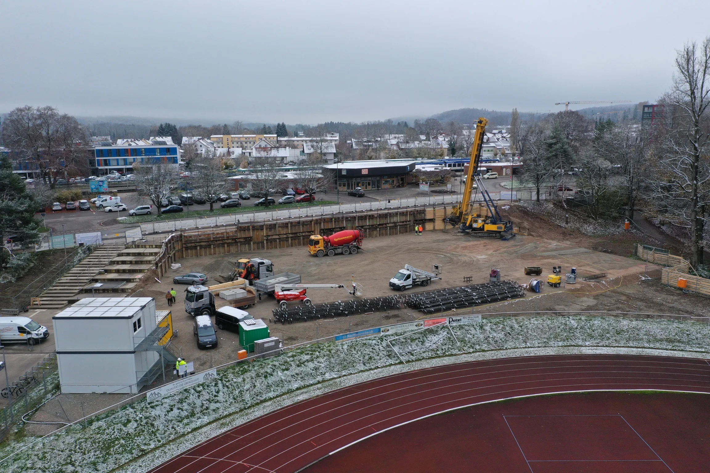 Baustelle mit Kran und Baucontainern neben einer Sportanlage mit roter Laufbahn.