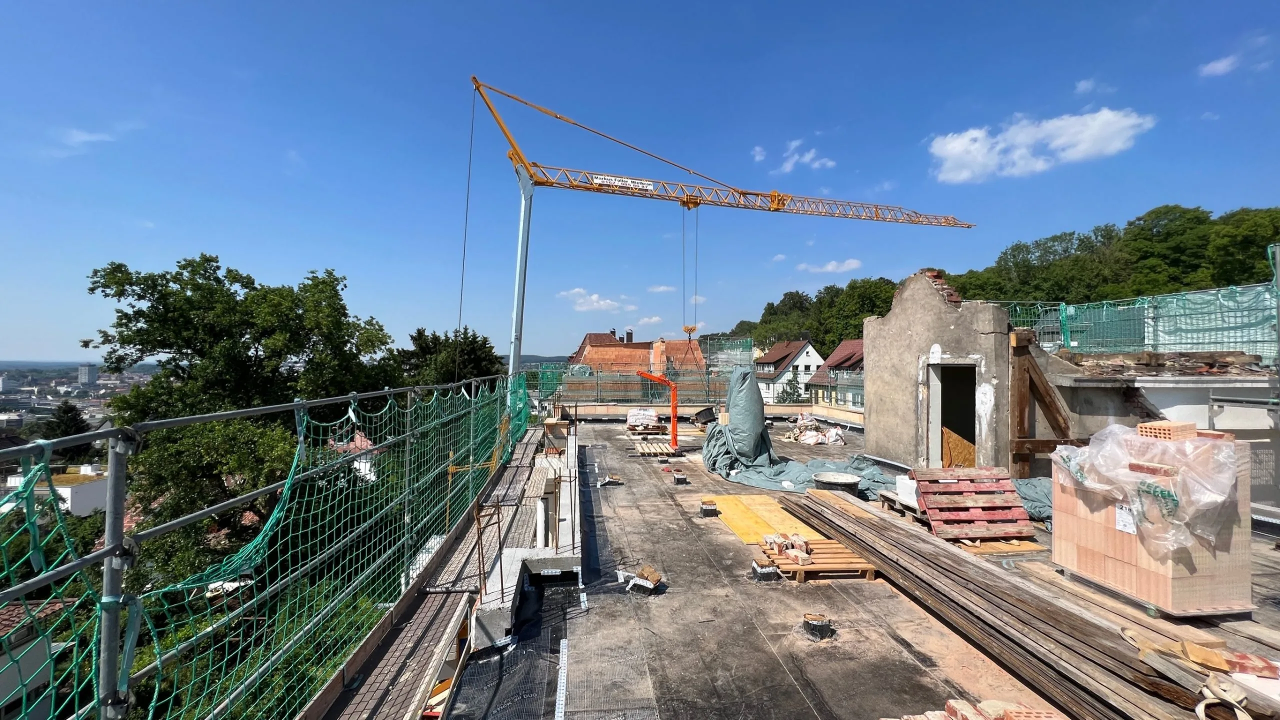 Baustelle im Bestand mit verbliebenen Mauerwänden, Baugerüst und Kran vor grünem Baumbestand.